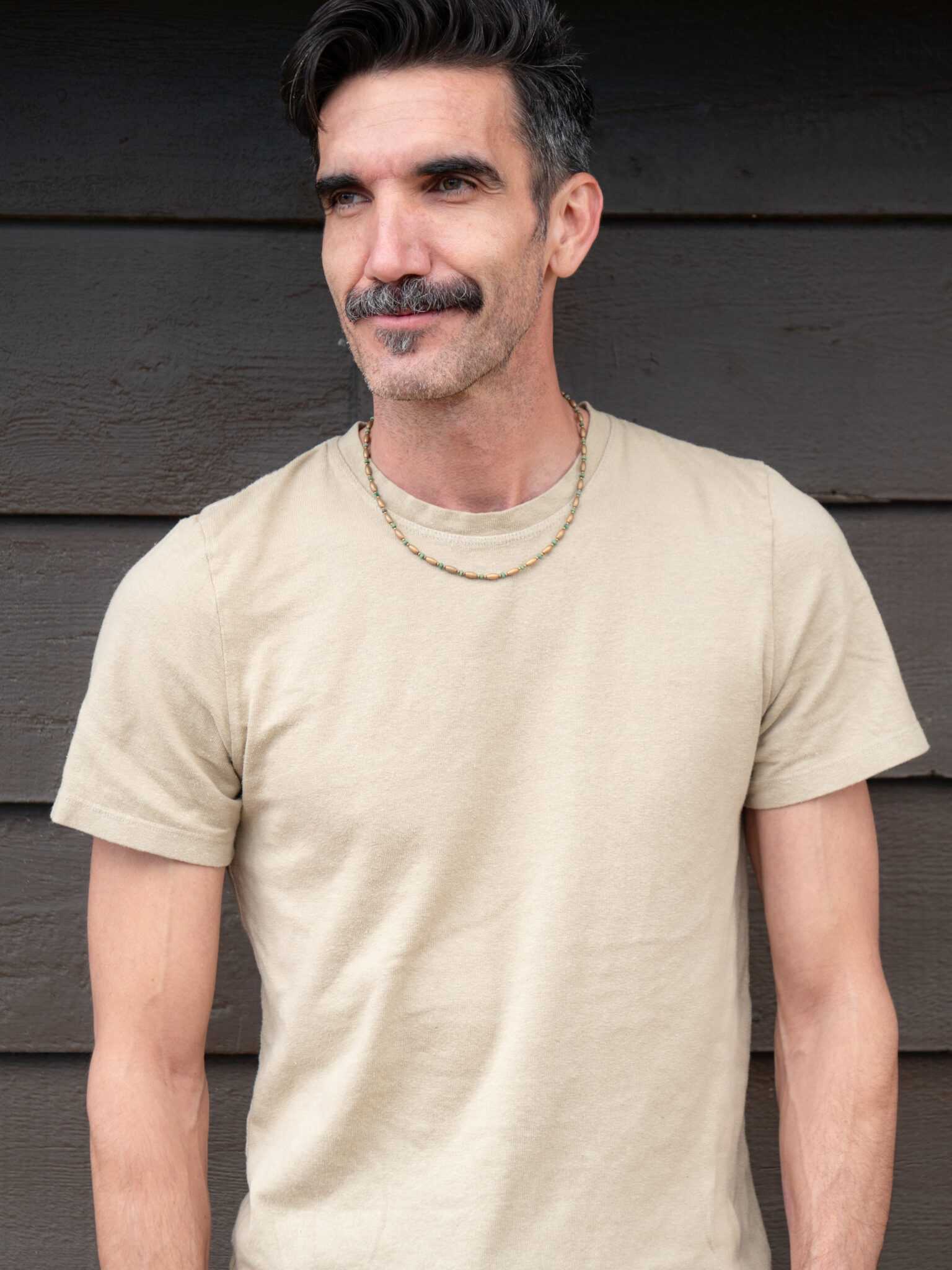 Man with short dark hair and a mustache wearing a beige t-shirt and two necklaces, standing in front of a dark wooden wall.