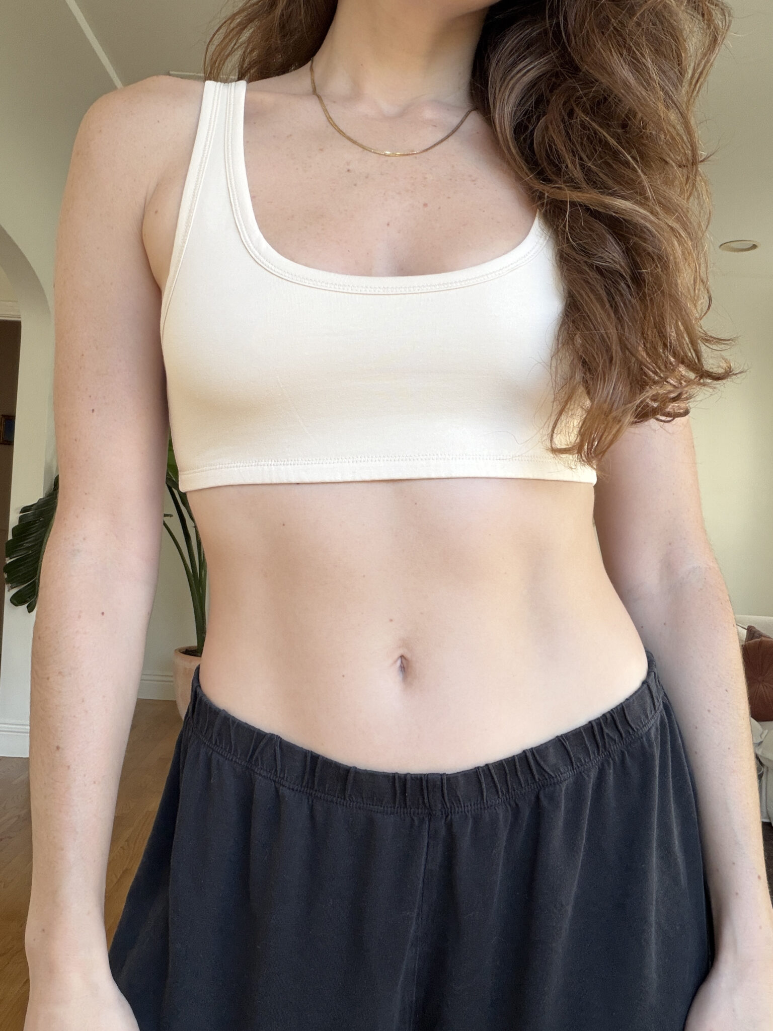 A person wearing a beige tank top and black pants stands indoors, with part of their face out of frame.