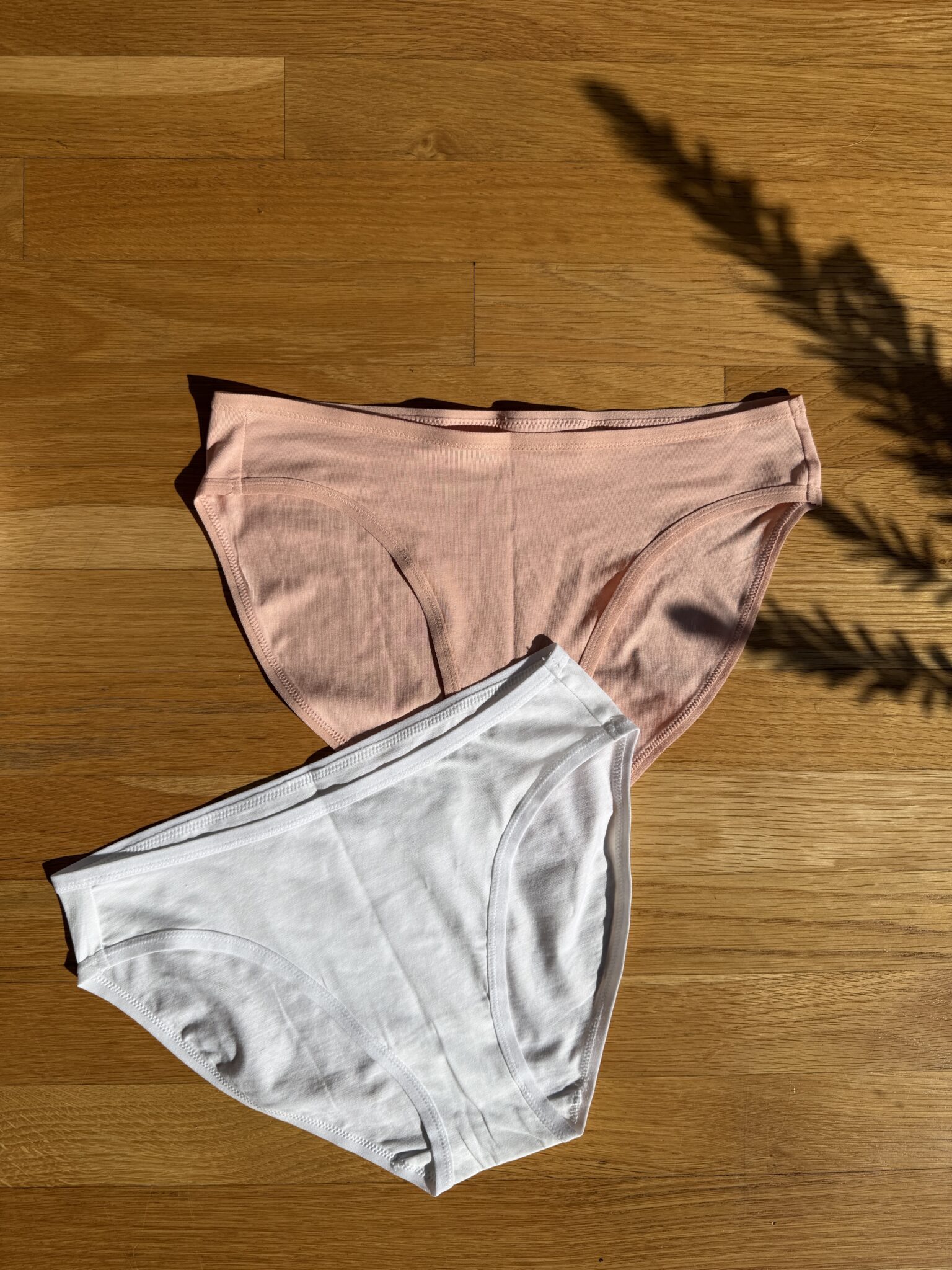 Two pairs of cotton underwear, one pink and one white, are laid flat on a wooden floor with a plant shadow visible on the right side.