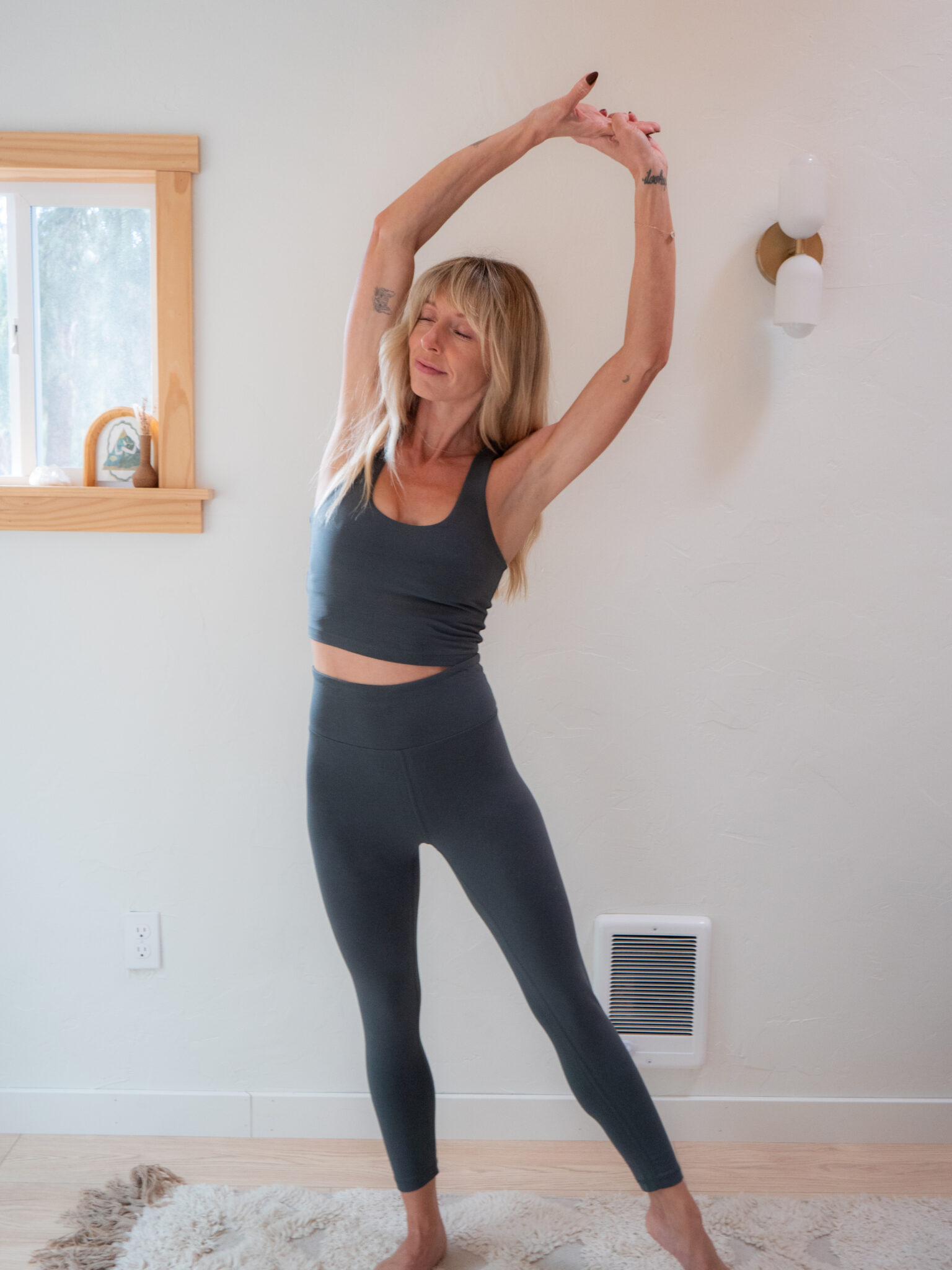 A woman in gray athletic wear stands indoors, stretching her arms overhead to one side.