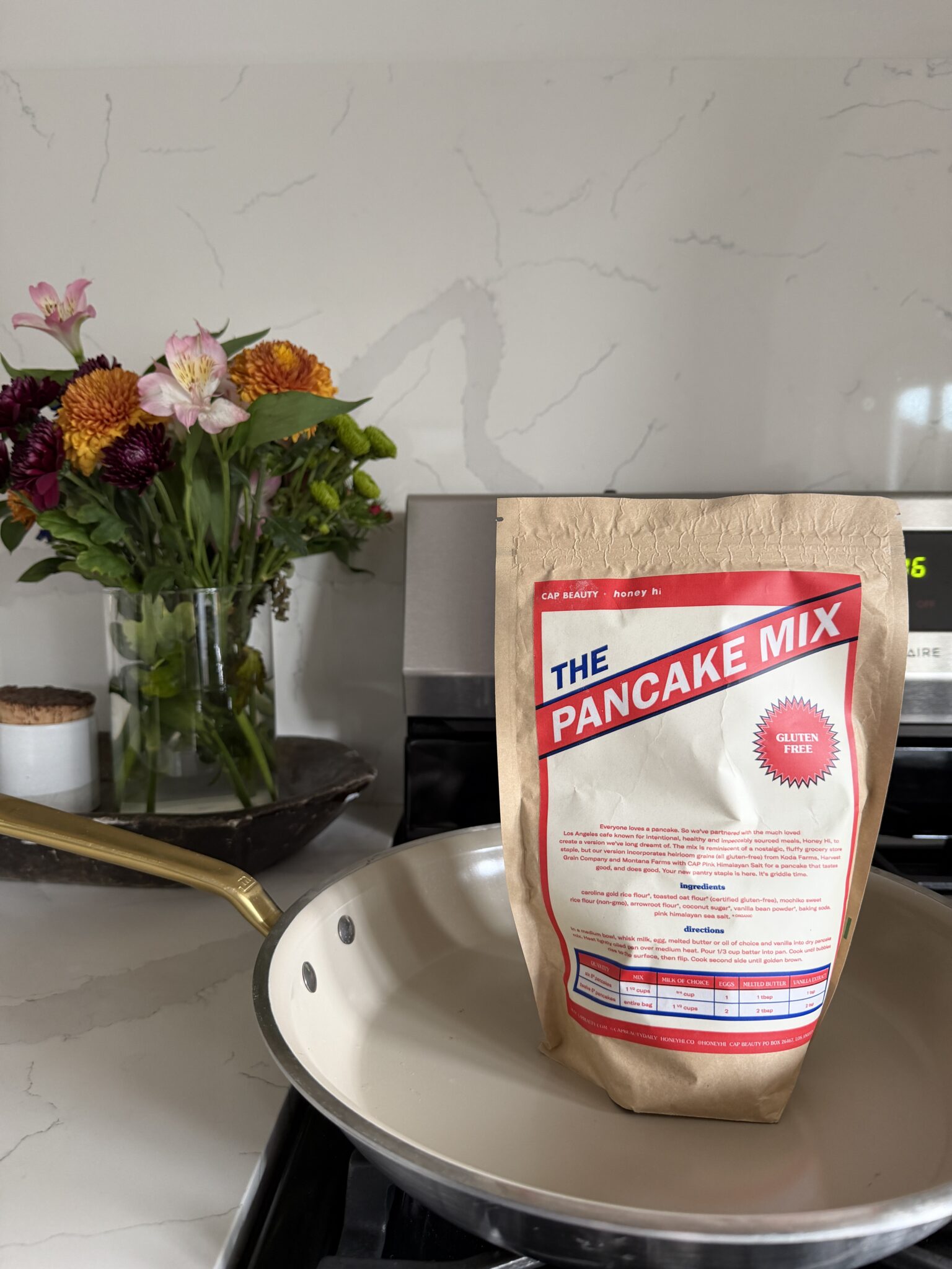 A bag labeled "The Pancake Mix" stands upright in a skillet on a stove, with a vase of flowers in the background.