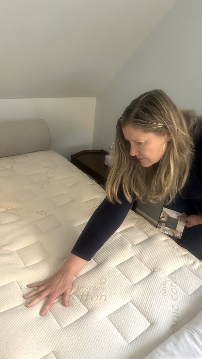A woman inspects a mattress by pressing her hand on it in a bedroom setting.