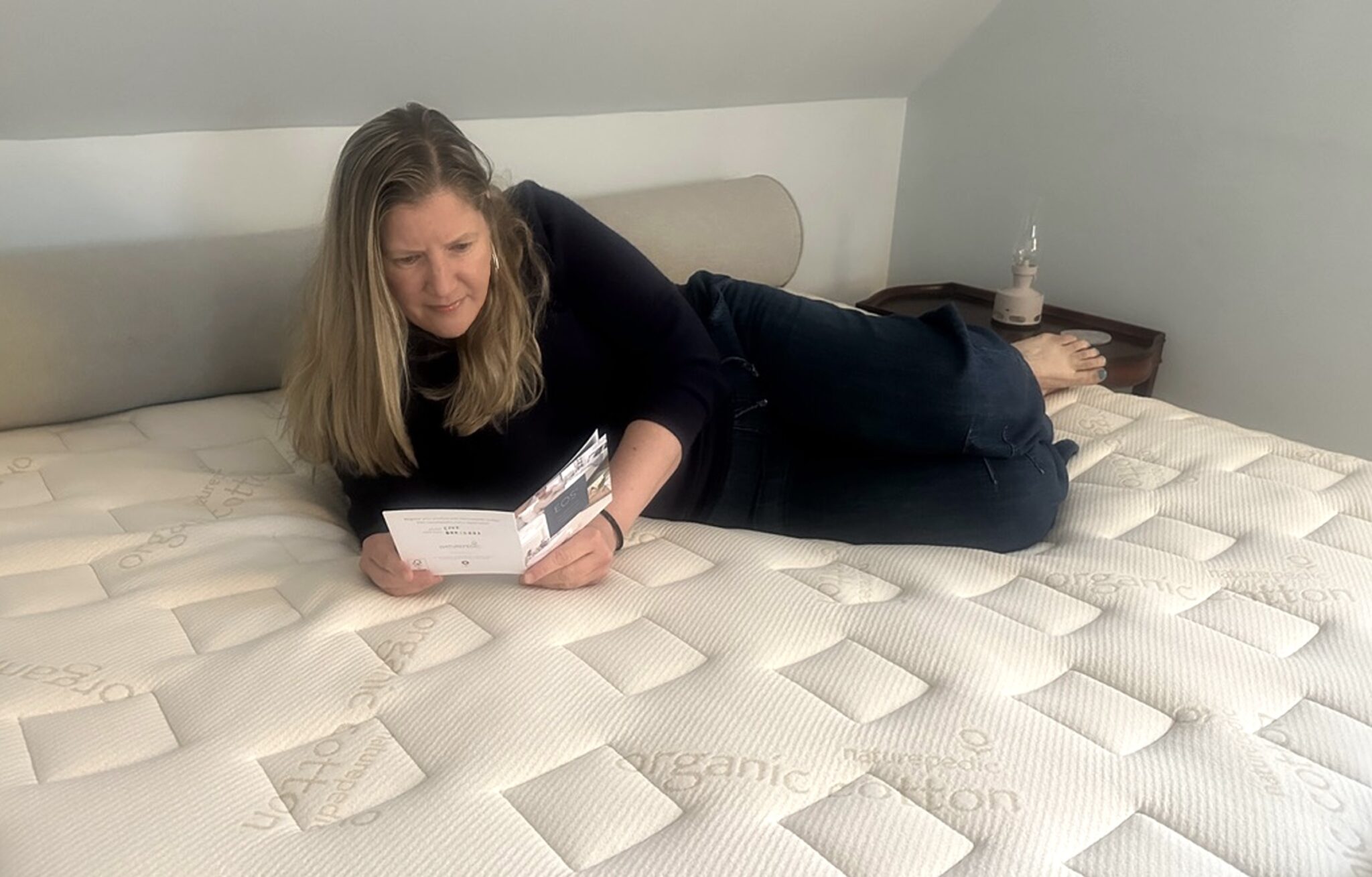 A woman lies on a bed reading a pamphlet. The mattress surface is labeled "organic cotton." A nightstand with a lamp is nearby.