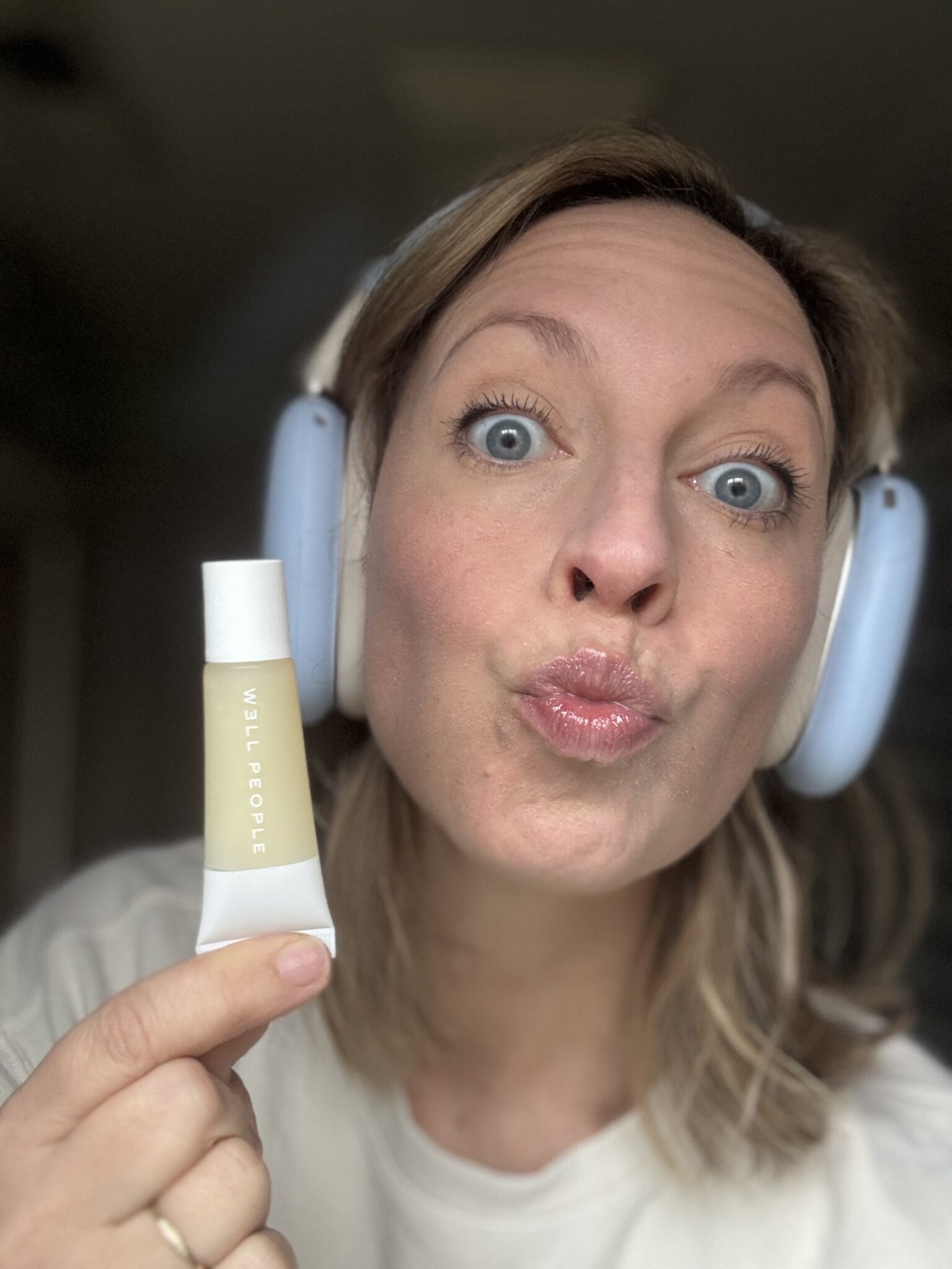 A person wearing over-ear headphones holds up a tube of W3LL PEOPLE cosmetic product and makes a puckered-lip face.