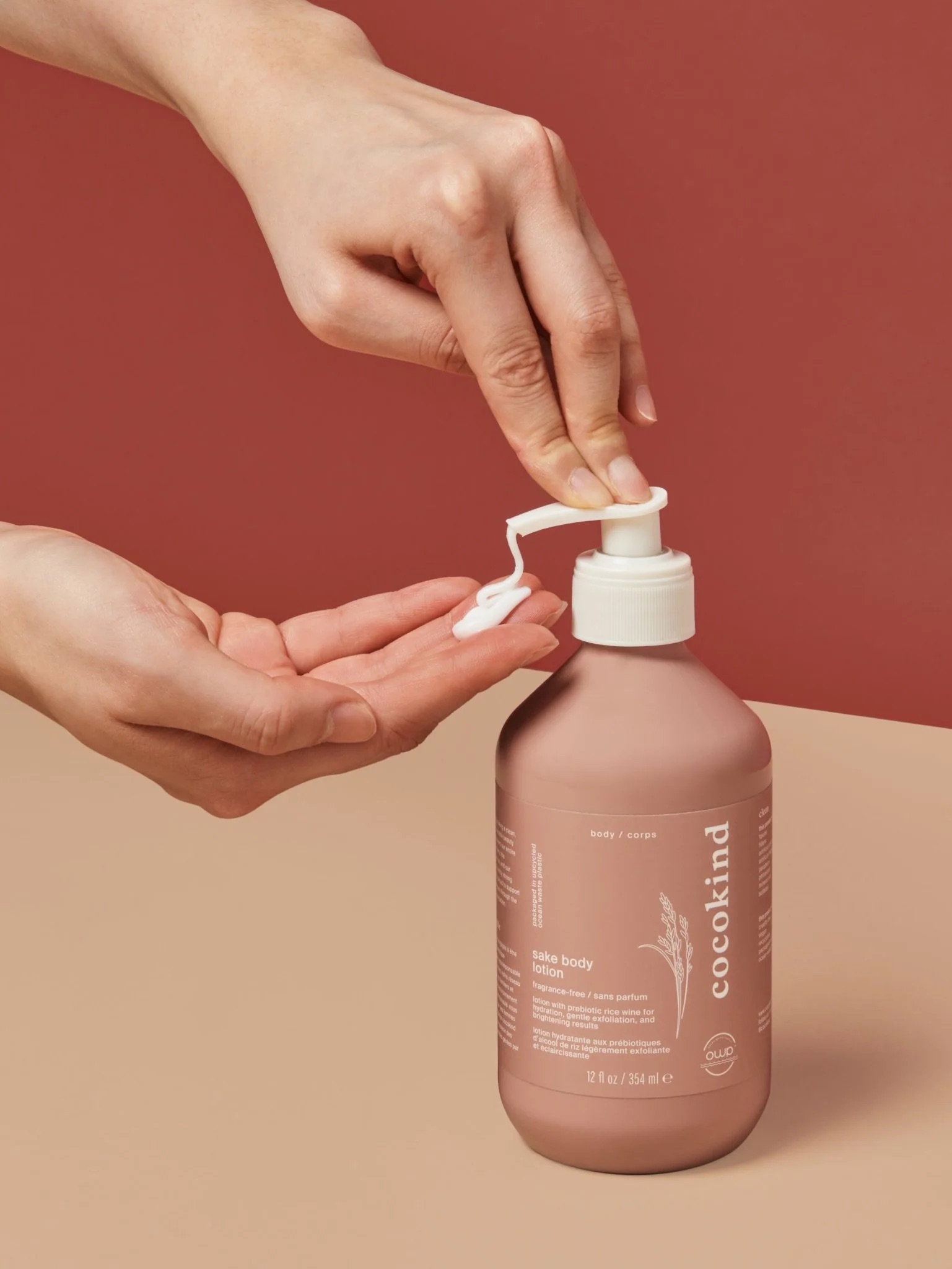A person dispenses lotion from a pink bottle labeled "cocokind" with one hand while holding the pump with the other hand against a red and beige background.