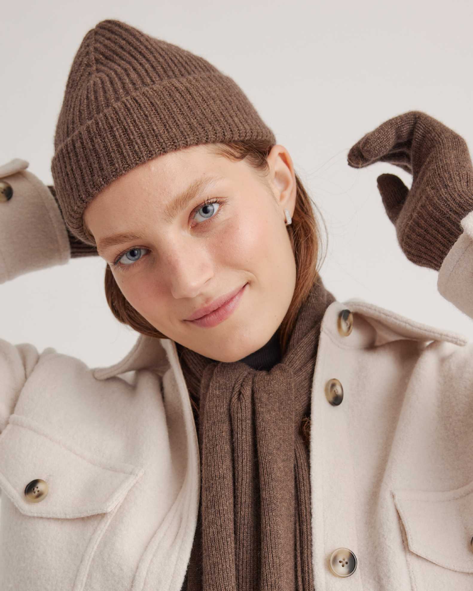 A person wearing a brown knit hat, matching scarf and gloves, and a beige button-up jacket, looking at the camera with a slight smile.