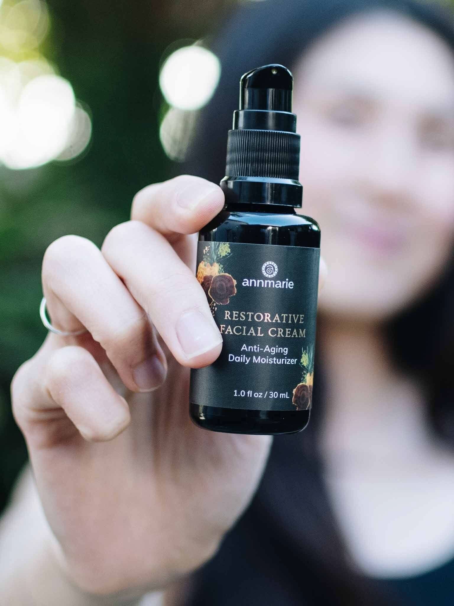 A person holds a bottle of Annmarie Restorative Facial Cream with a blurred natural background.