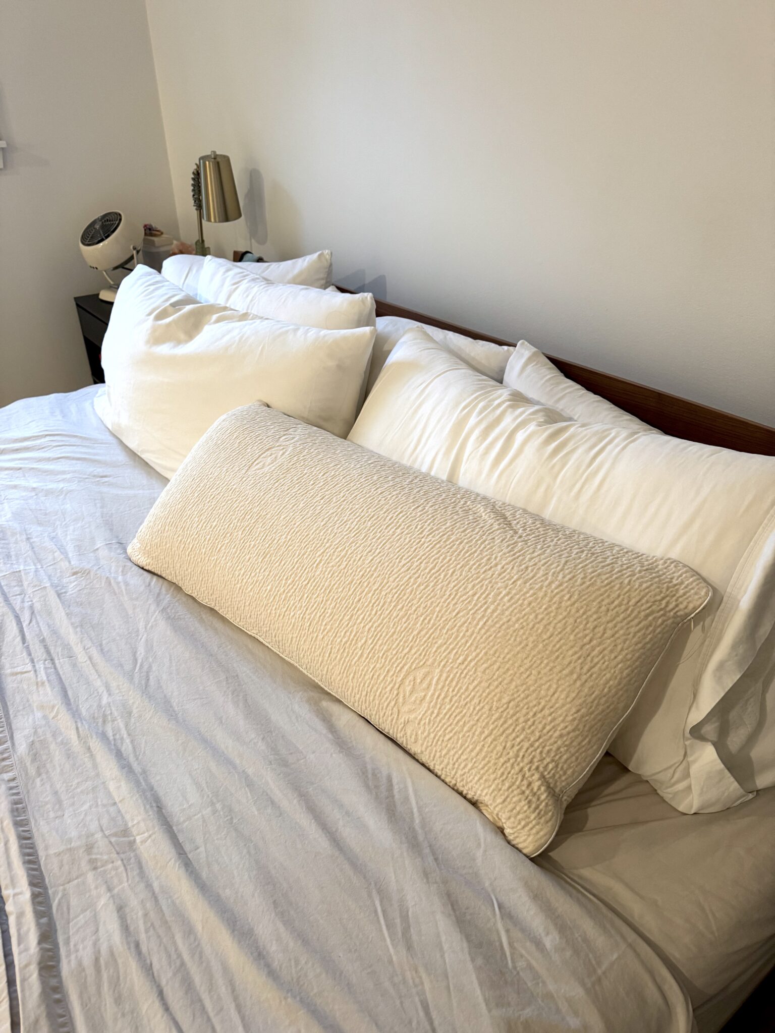 A neatly made bed with white pillows, a beige textured bolster pillow, and a nightstand with a lamp and clock in the corner.