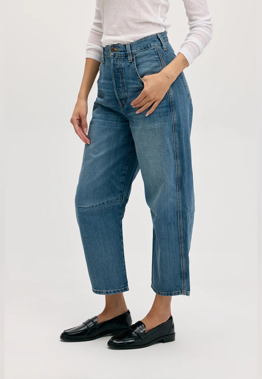 Person wearing high-waisted, relaxed-fit blue jeans, a white long-sleeve top, and black loafers, standing against a plain light background.