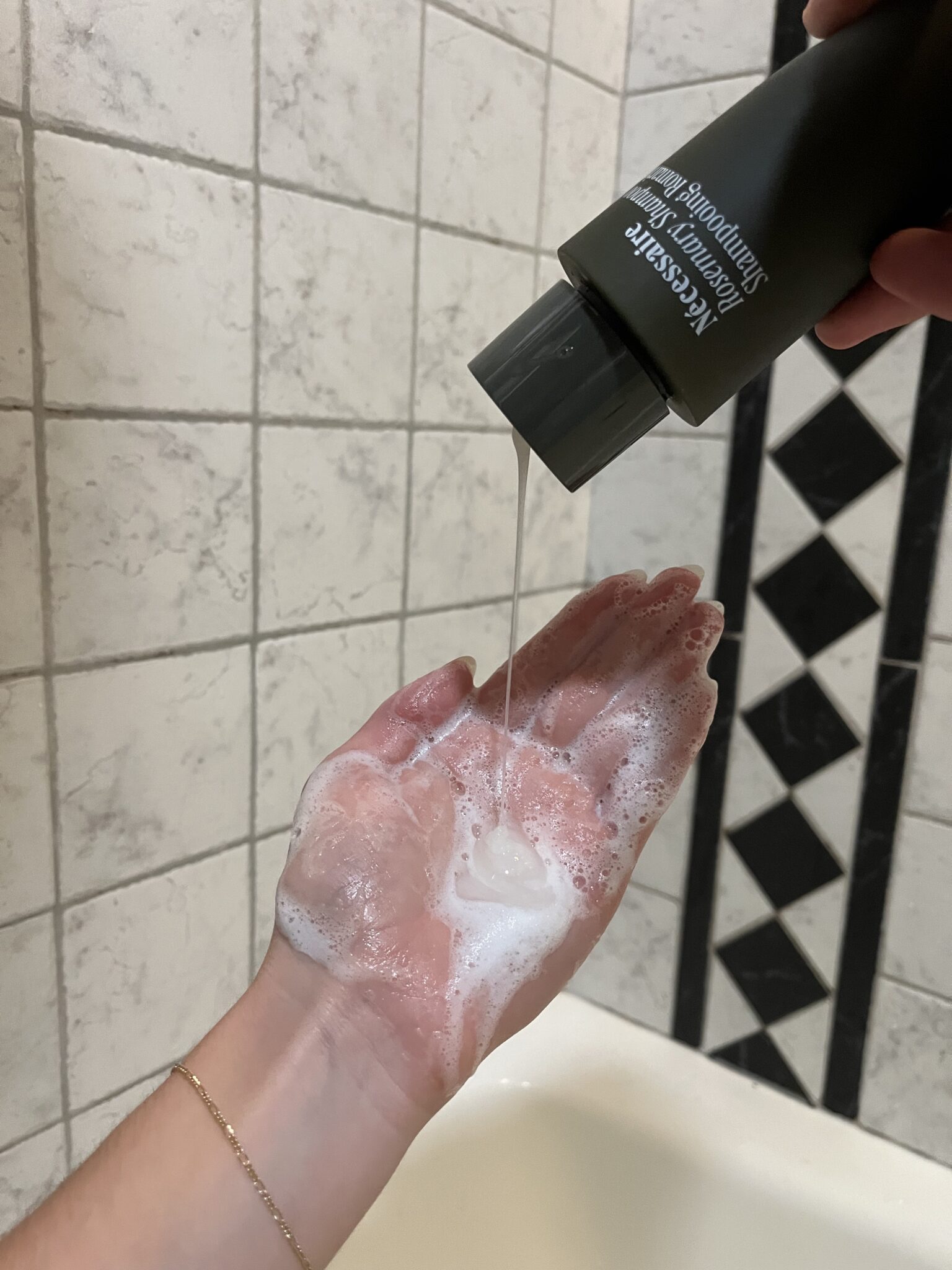 A person pours shampoo onto their soapy hand in front of a tiled bathroom wall.