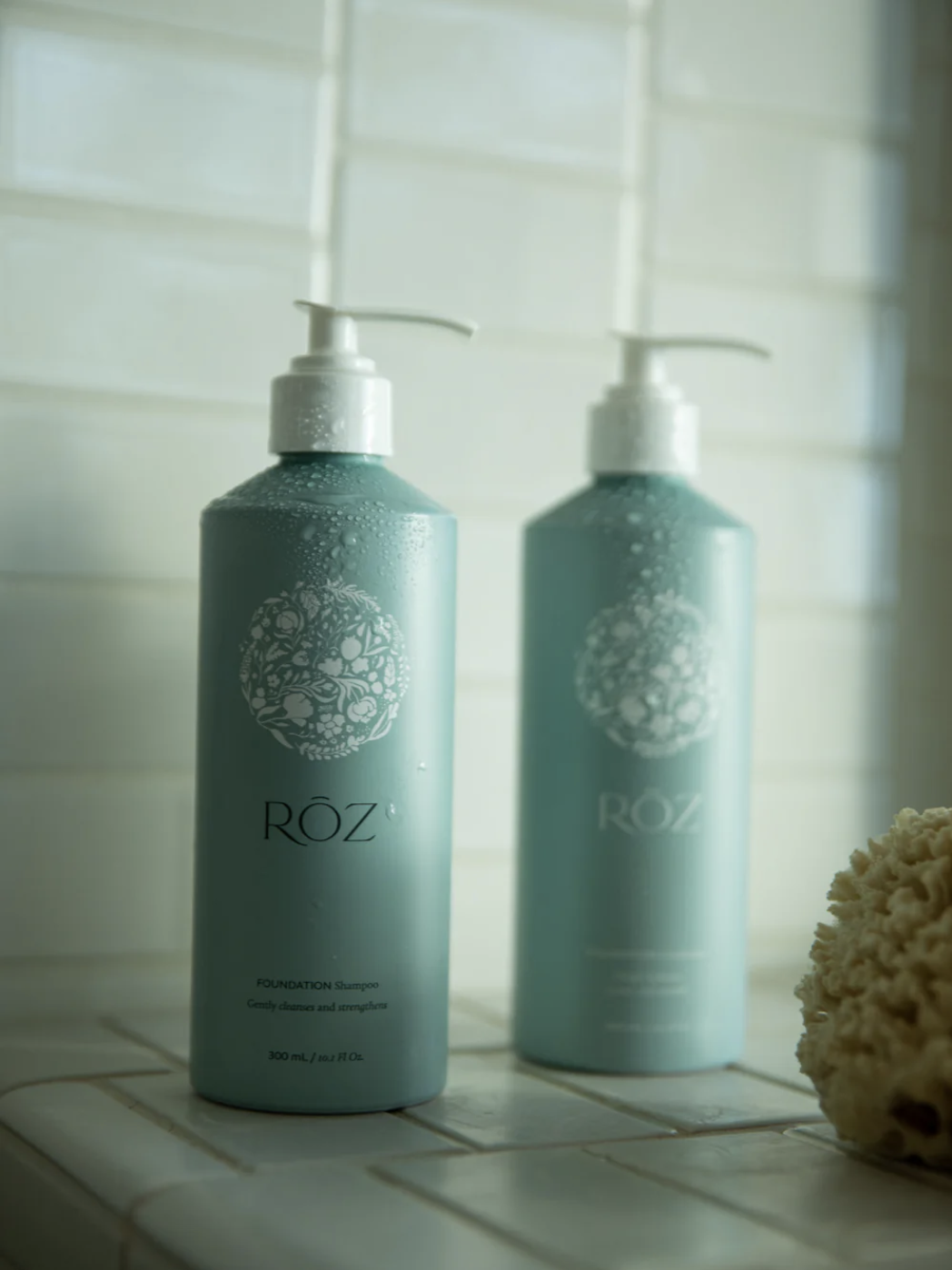 Two tall green RÓZ branded pump bottles, labeled "FOUNDATION Shampoo," standing on a tiled bathroom surface beside a natural sea sponge.