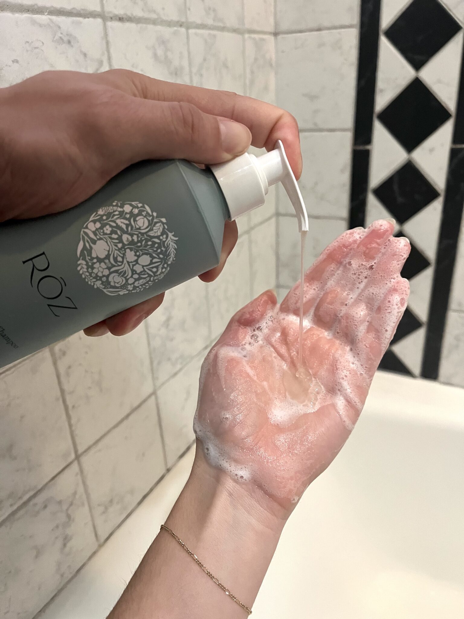 A person dispenses liquid from a ROZ bottle onto a soapy hand over a tiled bathroom surface.