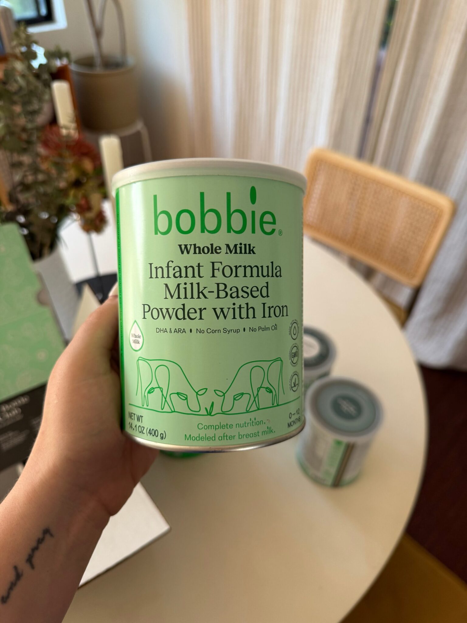 A hand holds a can of Bobbie Whole Milk Infant Formula Milk-Based Powder with Iron; two more cans sit on a white table in the background.