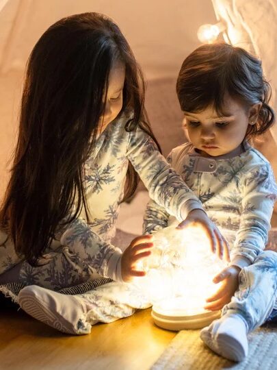 Two young children in pajamas sit inside a tent, touching a glowing light fixture placed on the floor between them.