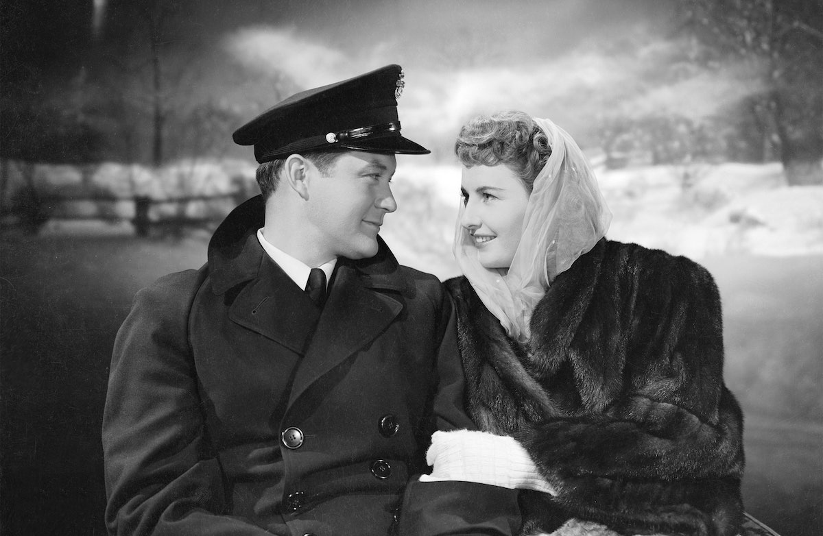 A man in a naval uniform and a woman in a fur coat and scarf sit close together, looking at each other and smiling in a winter setting.