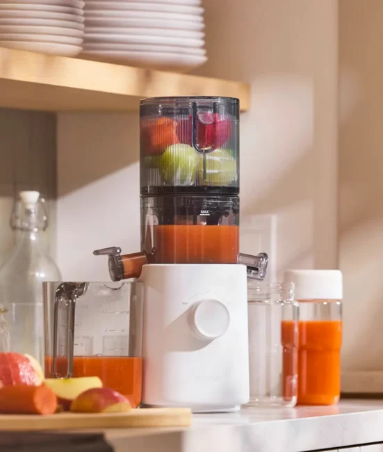 A juicer processes whole apples and other fruits, dispensing fresh juice into a container on a kitchen counter with jars, bottles, and cut fruit nearby.
