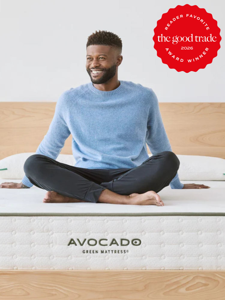 A man sits cross-legged on an Avocado Green Mattress in a bedroom. A red badge in the corner reads, "The Good Trade Reader Favorite Award Winner 2026.