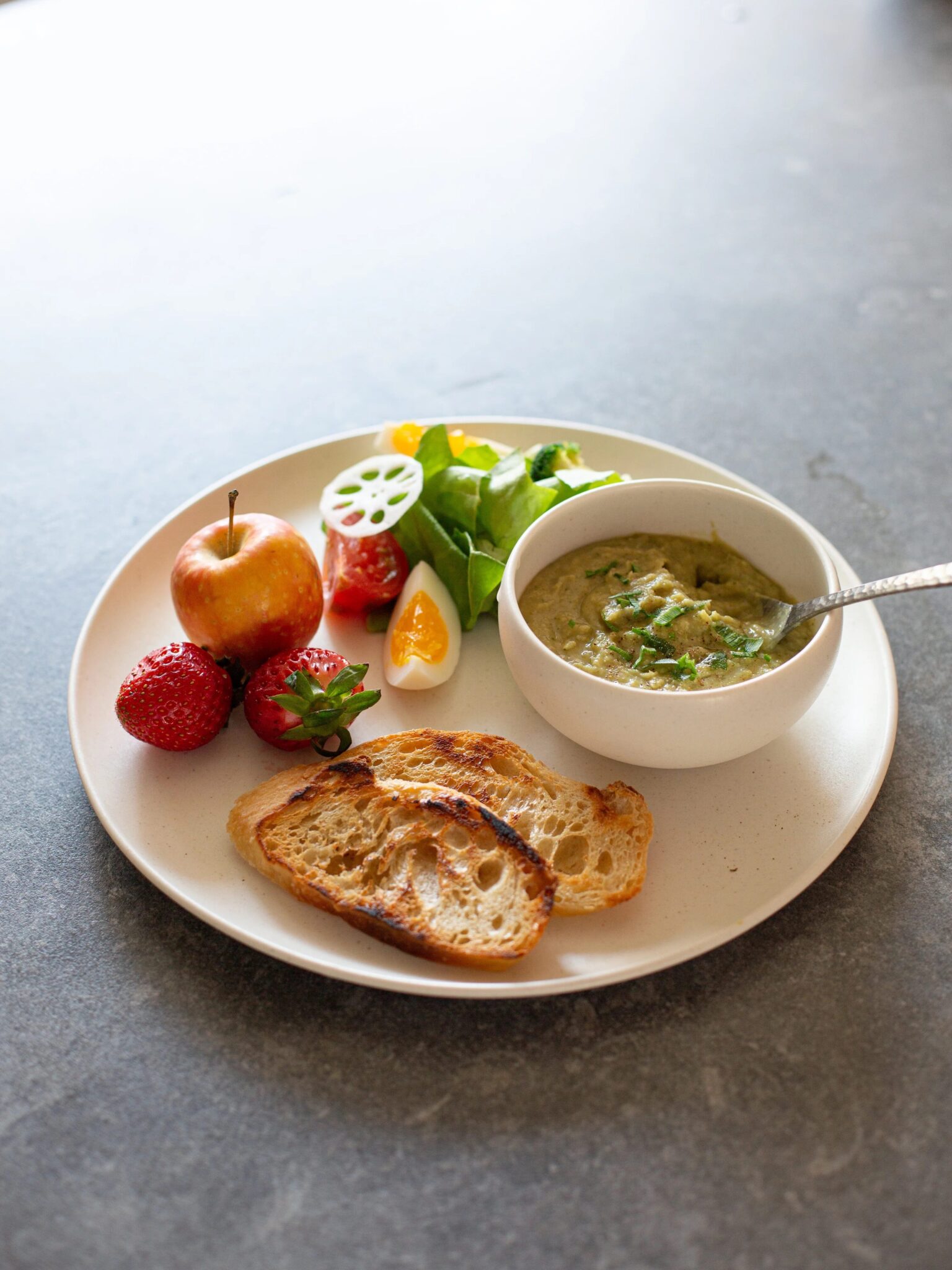 A plate with toasted bread slices, a bowl of soup or dip, an apple, strawberries, salad greens, sliced tomato, and a hard-boiled egg wedge on a gray surface.