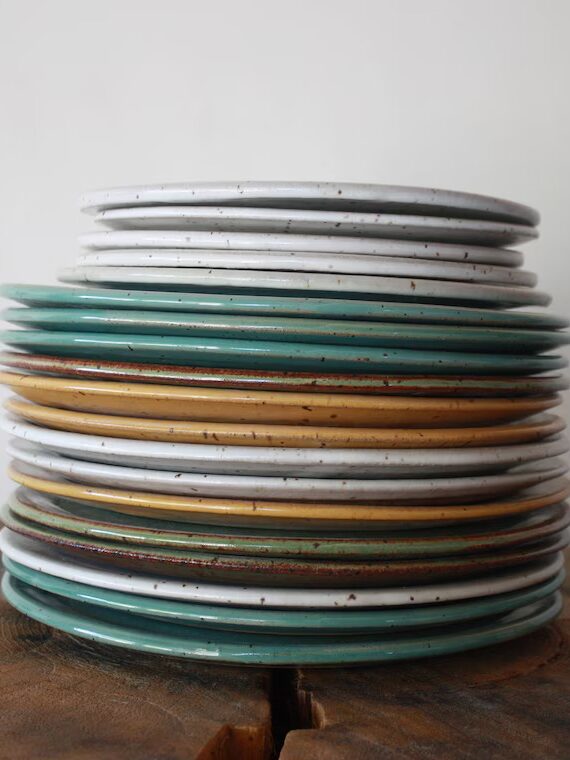 A stack of ceramic plates in white, green, mustard yellow, and brown colors sits on a wooden surface against a plain white background.