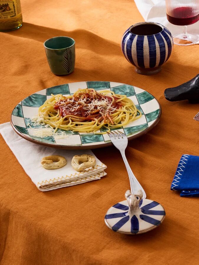 A plate of spaghetti with sauce and cheese sits on a checkered plate surrounded by a fork, bread, butter, cups, and ceramics on an orange tablecloth.