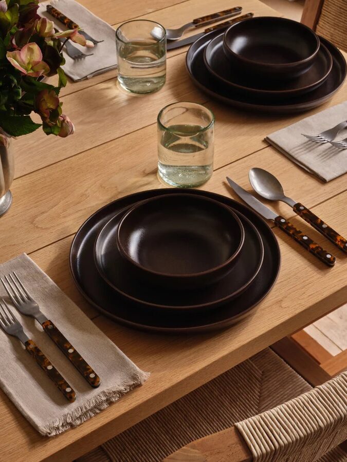 A wooden dining table set with dark brown plates, bowls, glasses of water, silverware with brown handles, cloth napkins, and a vase of flowers.