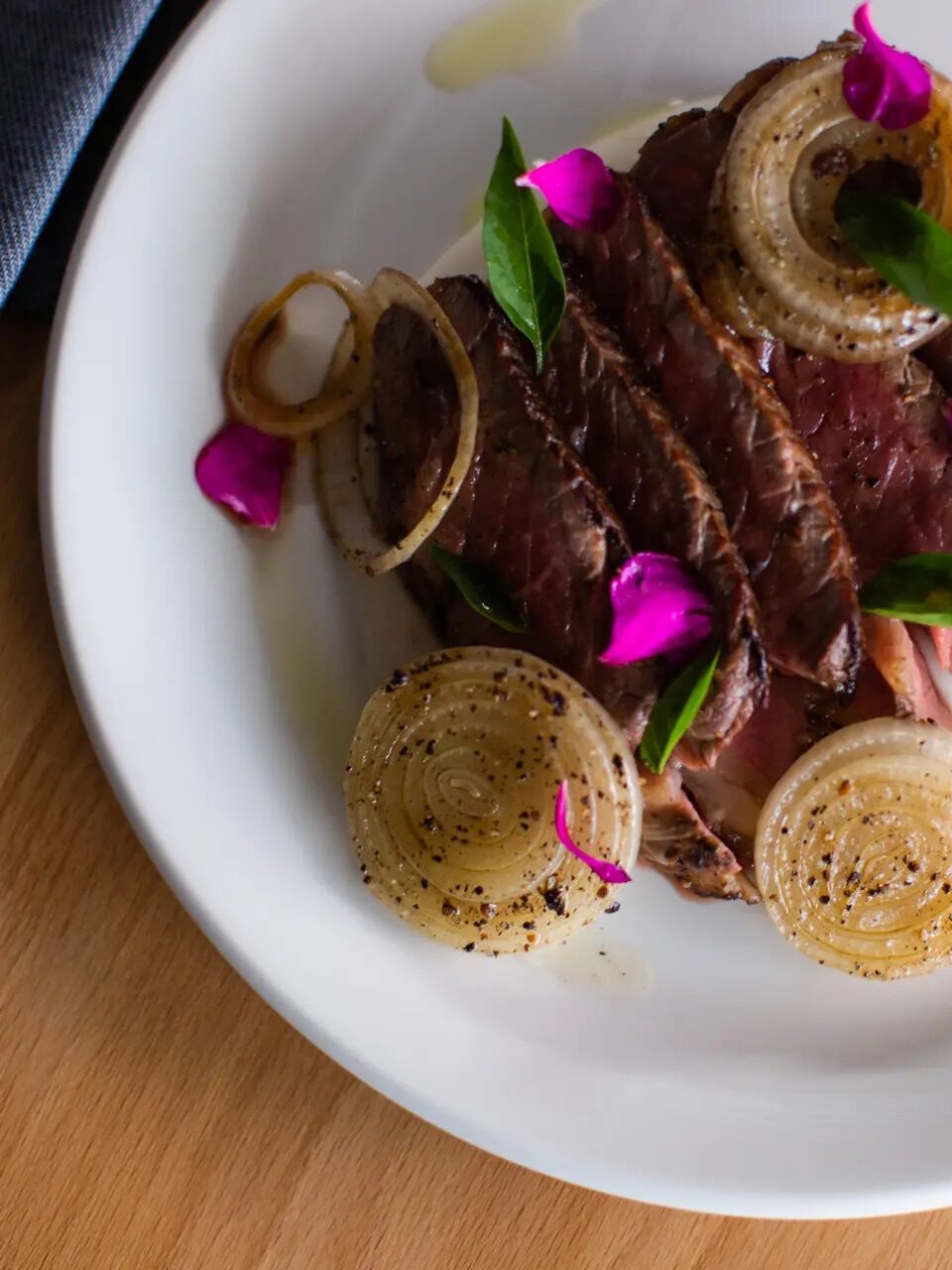 Sliced steak topped with grilled onion rings, green leaves, and purple flower petals, served on a white plate on a wooden table.