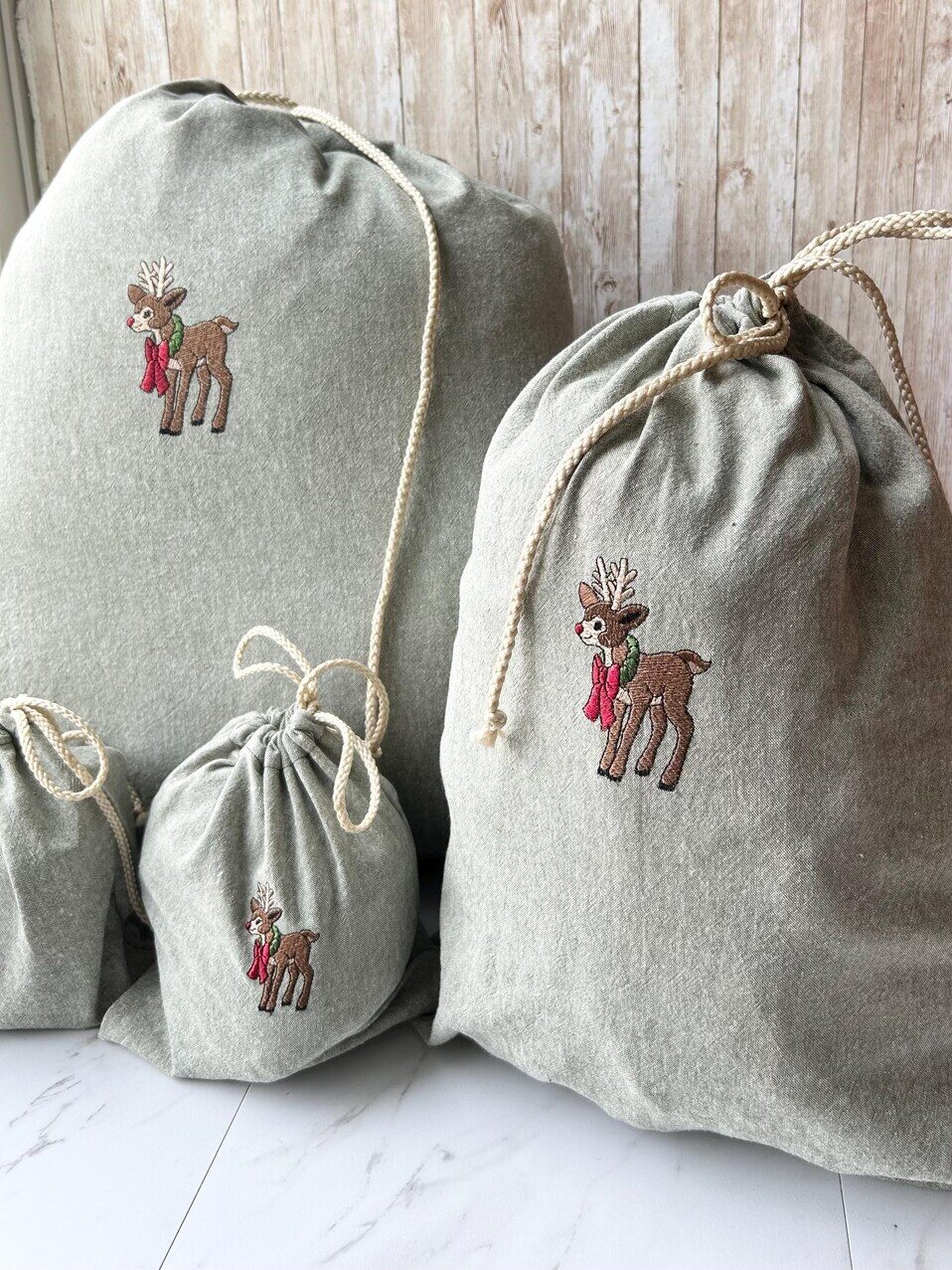 Three gray drawstring bags of varying sizes with embroidered reindeer and red scarves are arranged beside a red lantern on a white surface with a wooden background.