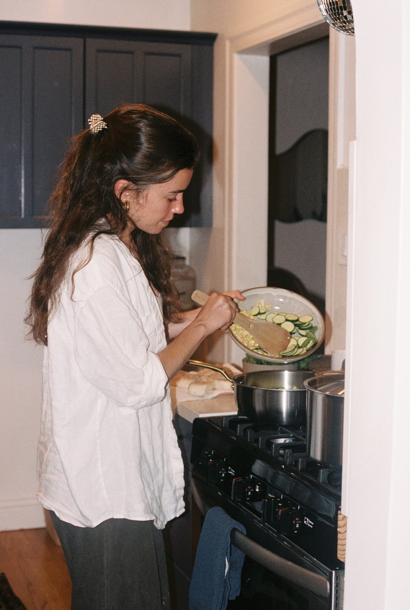 A woman stands at a stove, sautéing sliced zucchini in a pan in a kitchen.