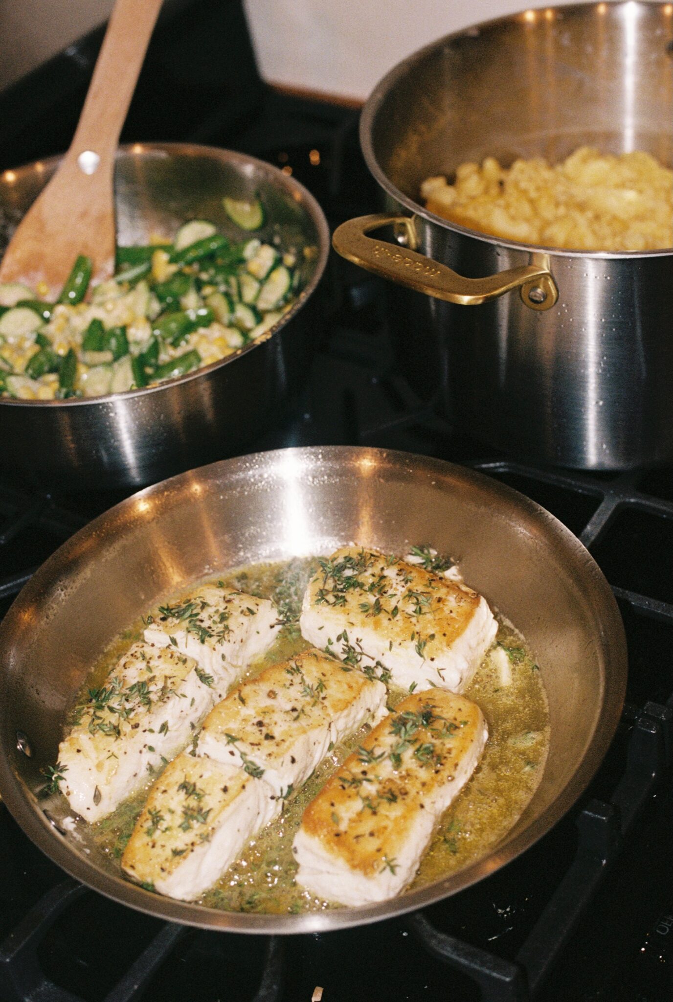 Four pieces of chicken breast cook in a skillet with herbs and sauce; a pot of pasta and a pan of sautéed vegetables are on the stove.