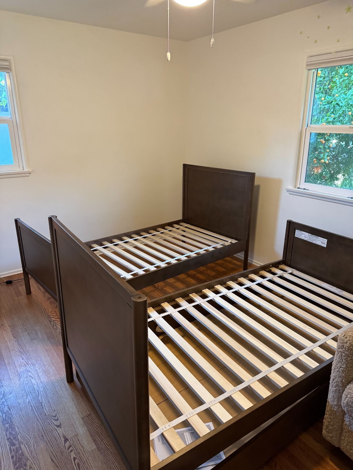 Two wooden twin bed frames with slatted bases are positioned side by side in a room with hardwood floors, white walls, and two windows showing greenery outside.