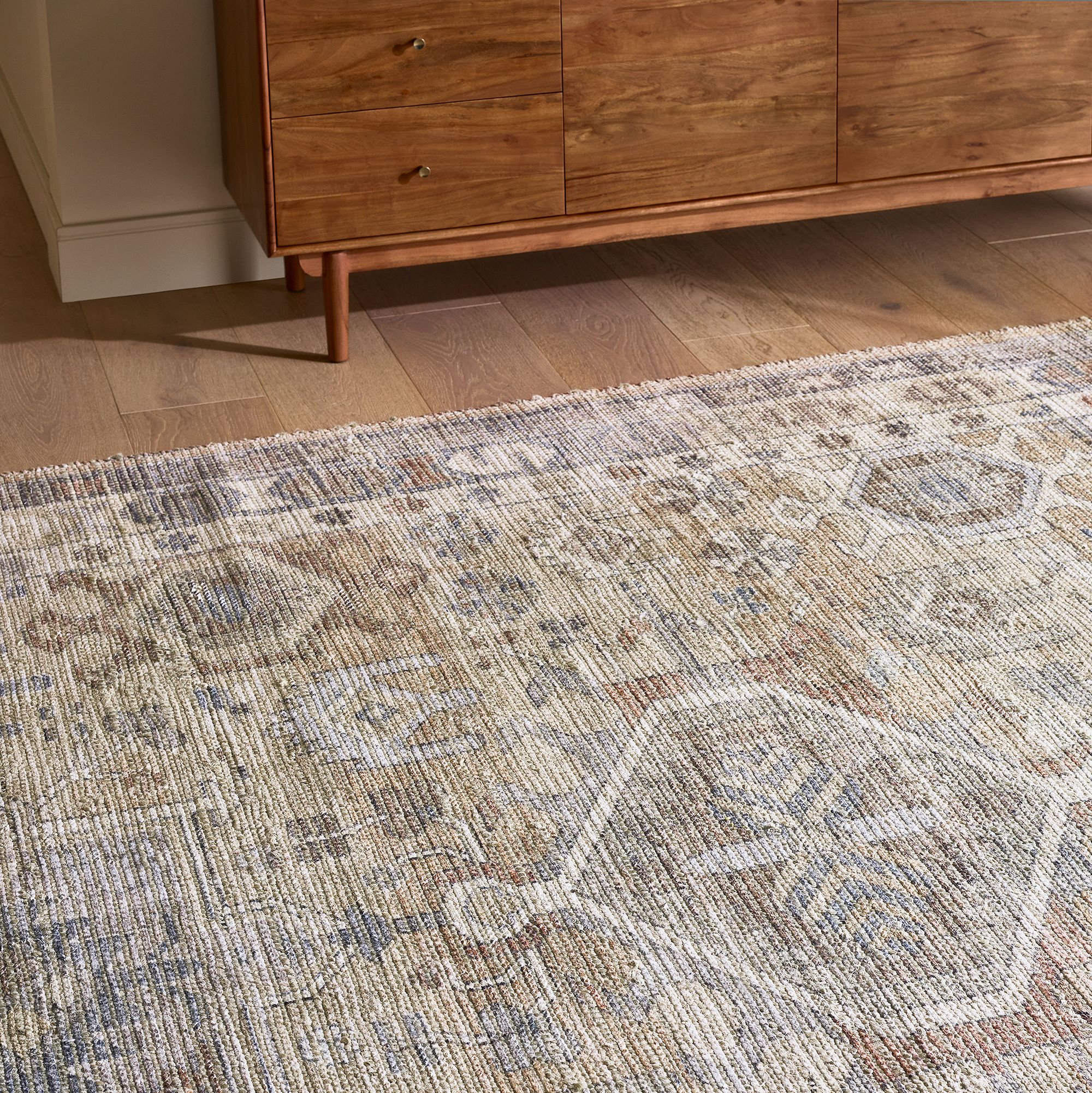 A patterned area rug with muted colors lies on a wooden floor in front of a wooden dresser with angled legs.
