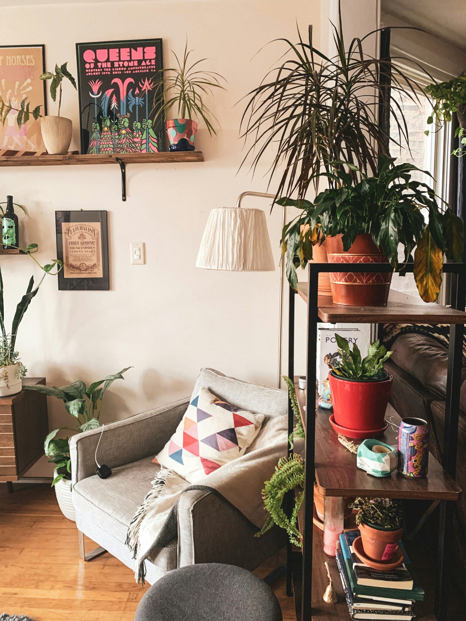 A cozy living room corner with a gray armchair, colorful geometric pillow, various potted plants, books, framed posters, and wooden shelves on a light wall.