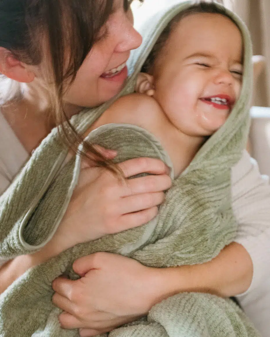 An adult smiles while holding a laughing child wrapped in a green towel.