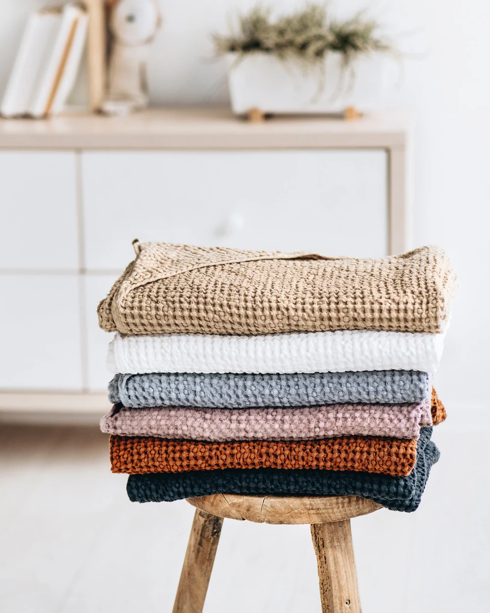 A stack of folded waffle-knit blankets in various colors sits on a wooden stool in a bright, minimalist room with a cabinet and plant in the background.