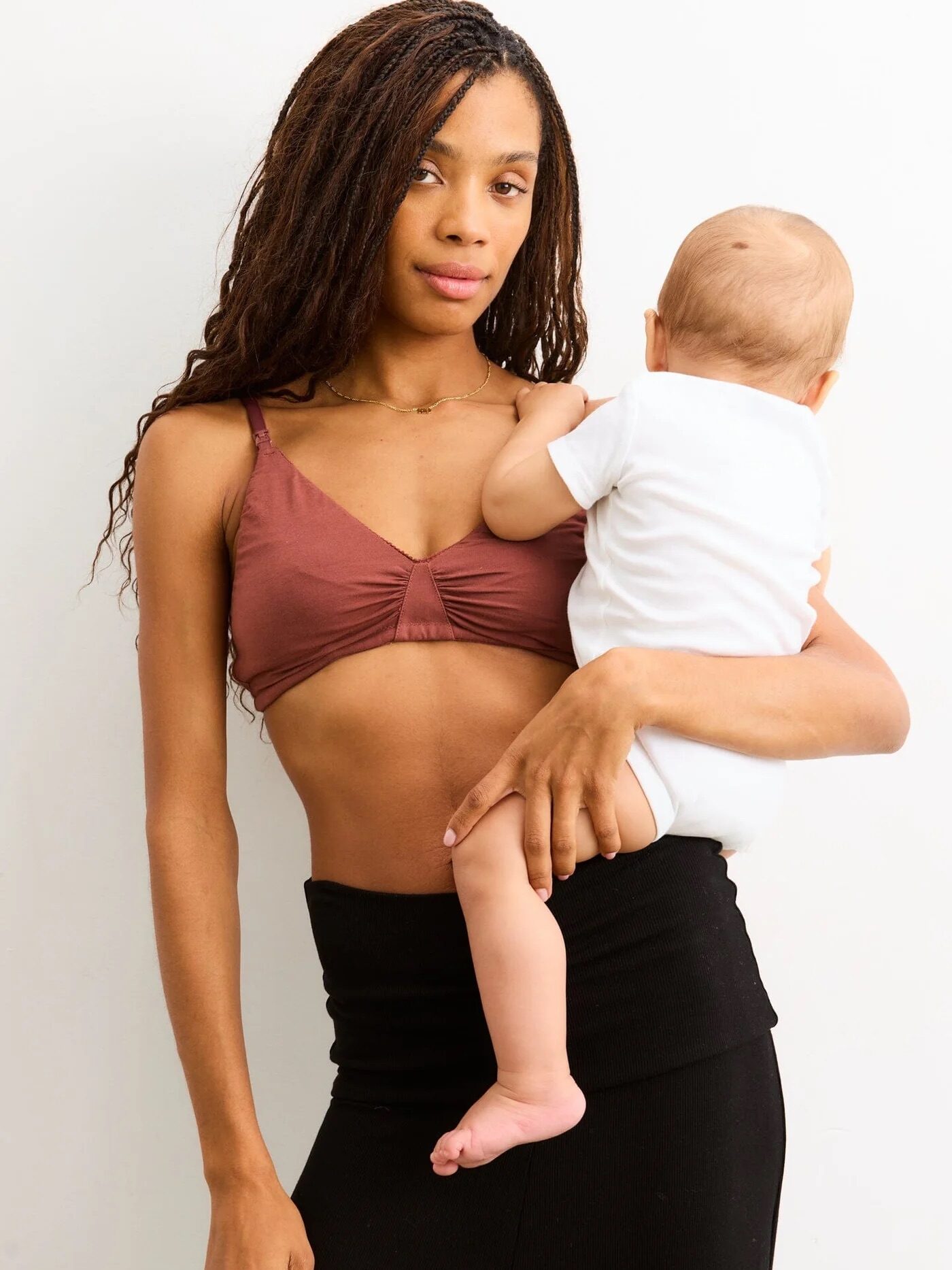 A woman in a brown bra and black skirt holds a baby dressed in a white onesie, standing against a plain white background.