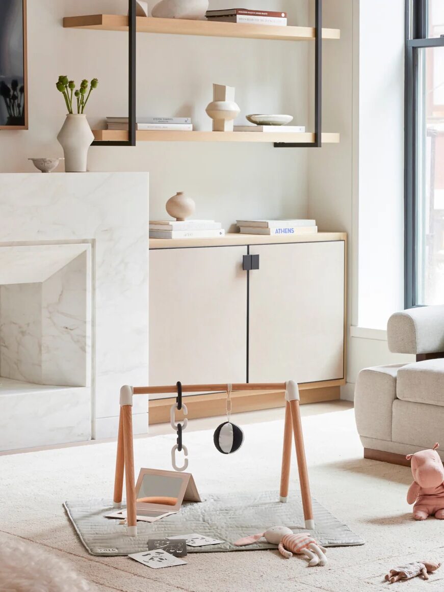 A modern living room with light-colored furniture, a marble fireplace, shelves with decor, and a baby play gym with toys on a soft rug.