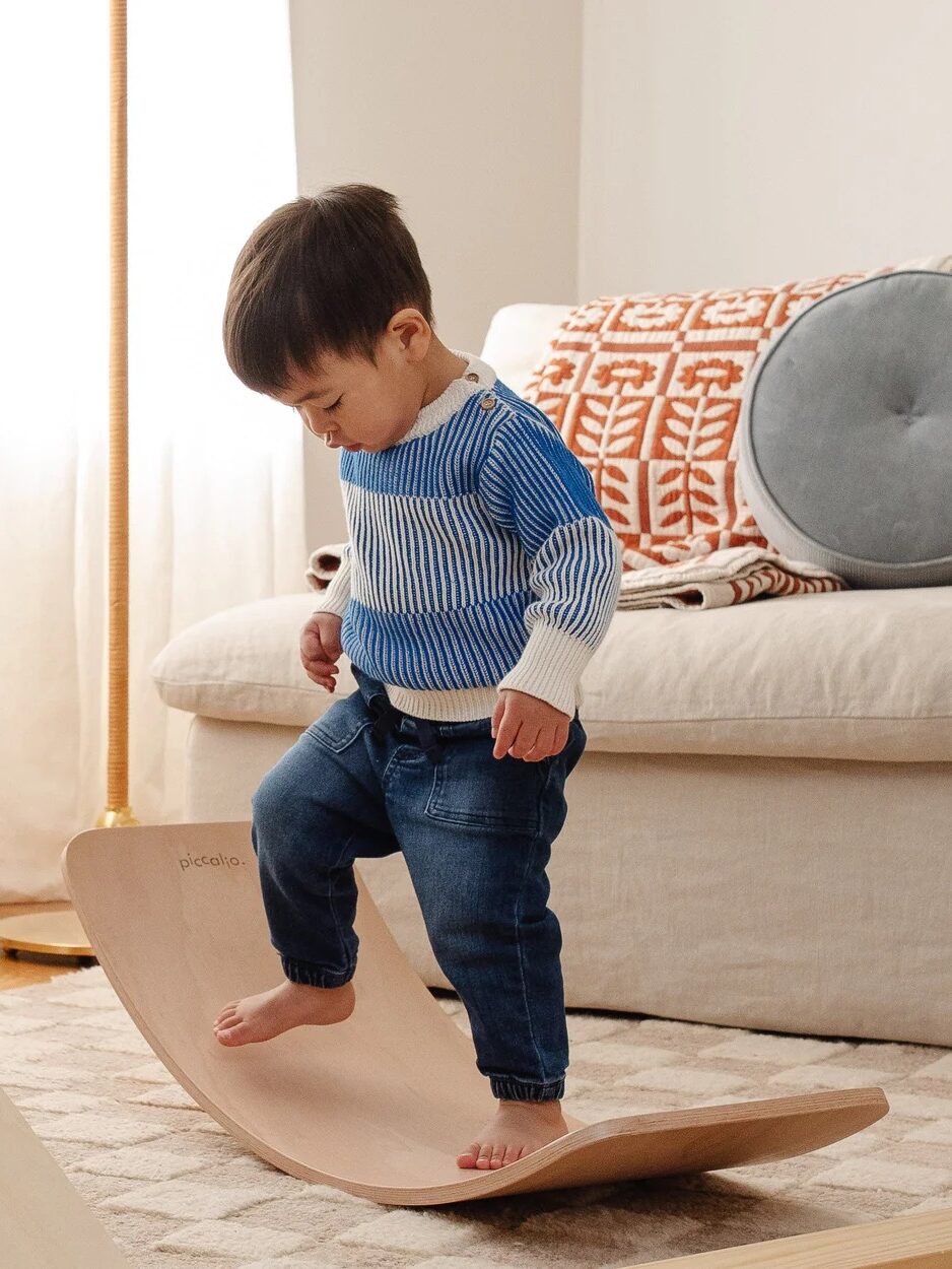 A young child balances barefoot on a curved wooden board in a living room, with a beige sofa and patterned cushion in the background.
