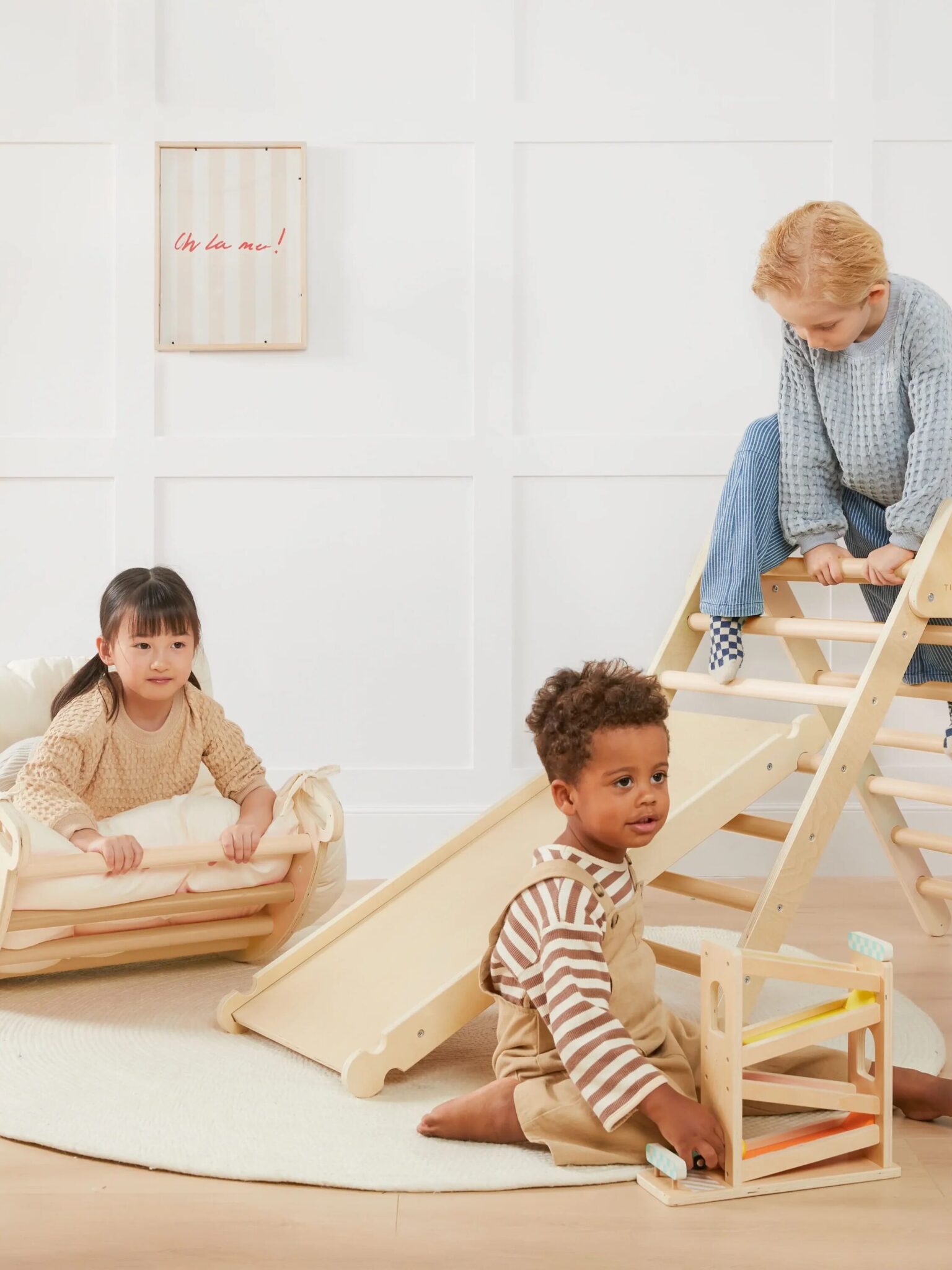 Three young children play indoors with wooden climbing, rocking, and sliding toys on a light rug against a white wall.