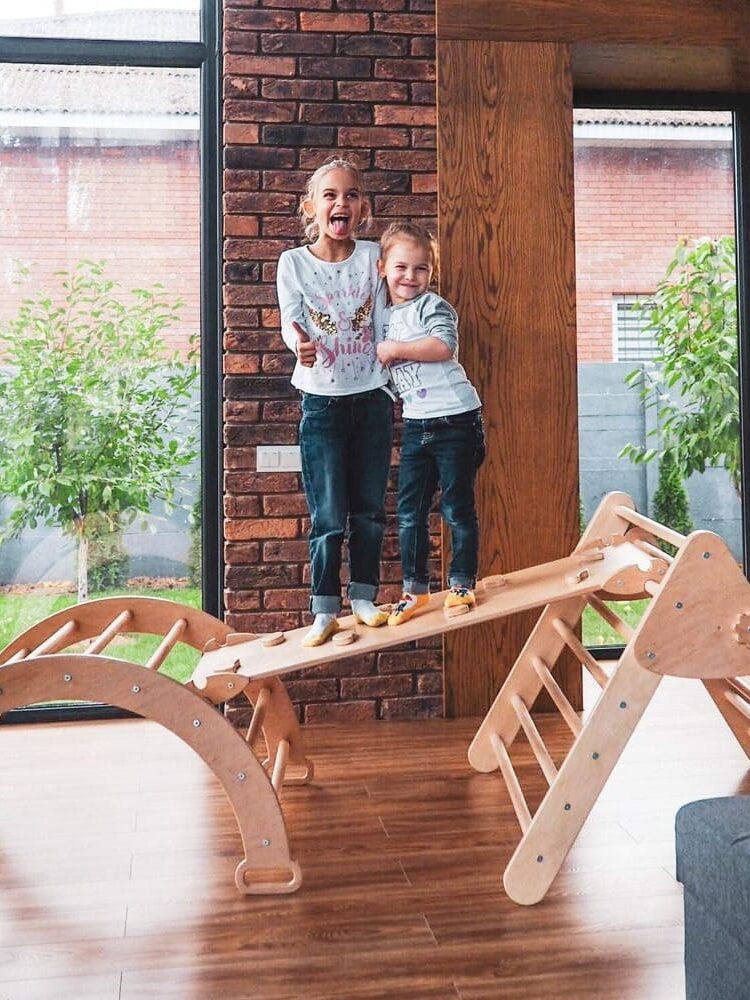 Two children stand and smile on a wooden climbing toy structure inside a living room with large windows and brick walls.