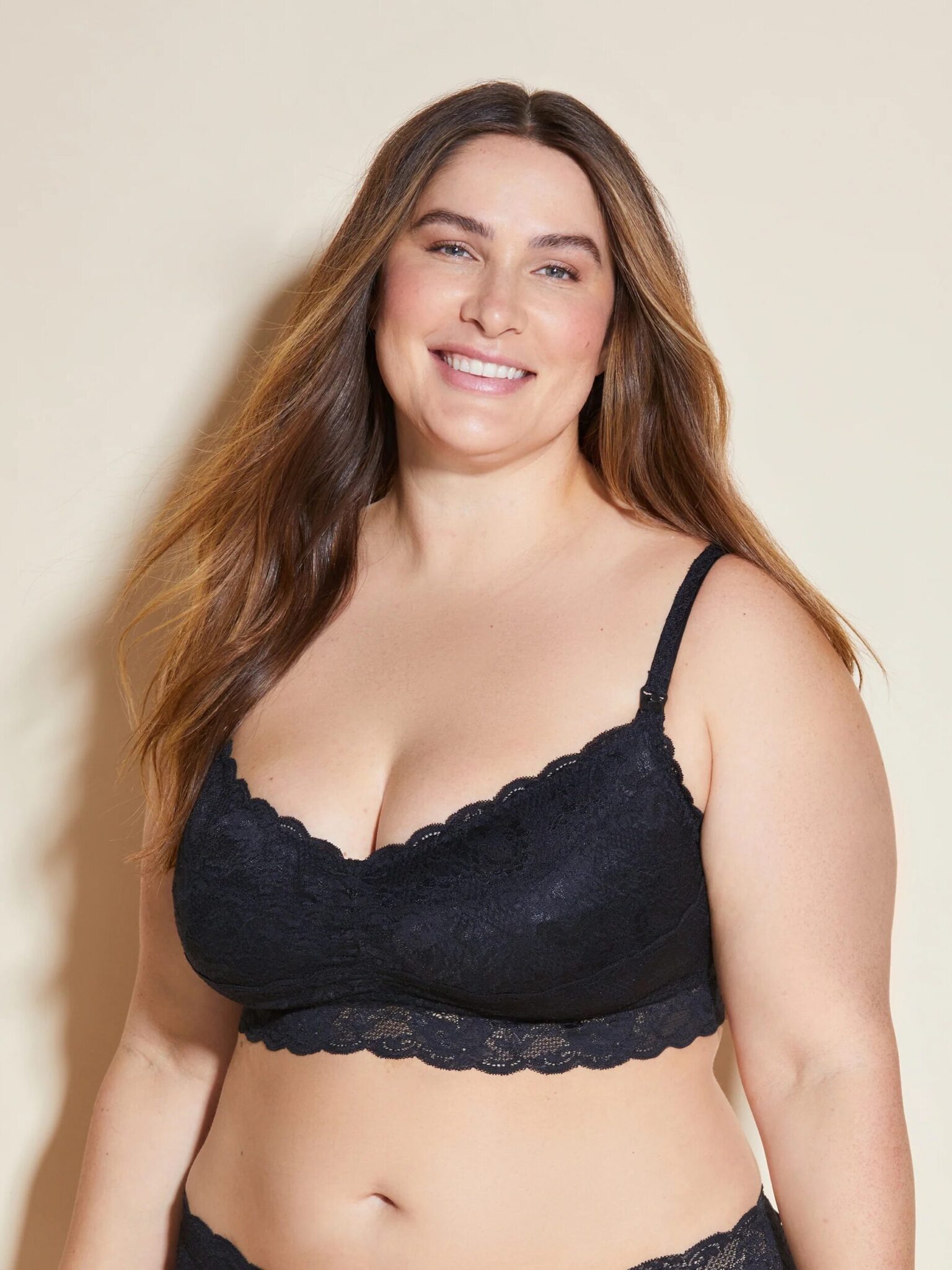A woman with long brown hair wears a black lace bralette and matching underwear, standing against a plain beige background and smiling.