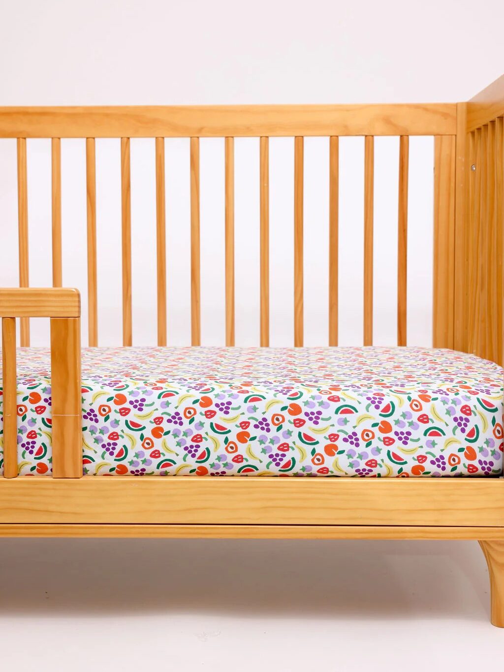 Wooden baby crib with vertical slats, featuring a mattress covered in a colorful, floral-patterned sheet against a plain white background.