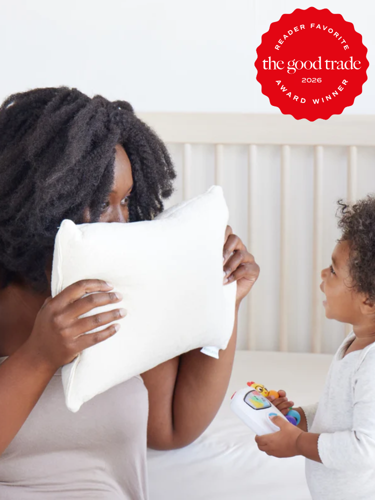 A woman holds a white pillow in front of her face while sitting on a bed next to a small child holding a toy. A red "The Good Trade 2026 Award Winner" badge appears in the top right corner.