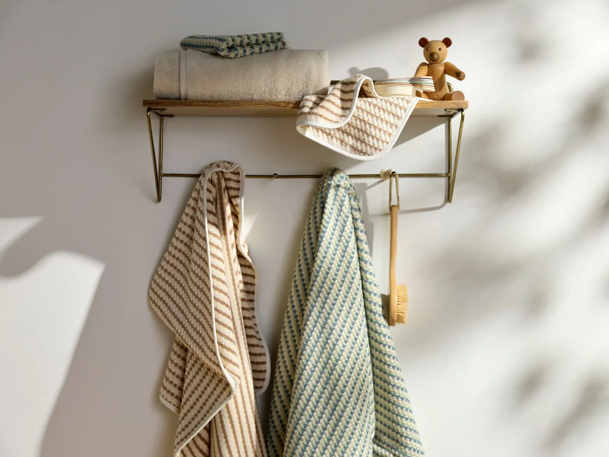A wooden wall shelf holds folded towels, a small teddy bear, and a brush, with two striped towels and a bath brush hanging below.