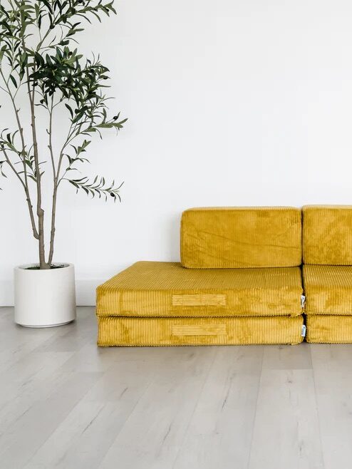A mustard yellow, modular floor sofa with two seat cushions and two backrests sits against a white wall next to a potted plant on a light wood floor.