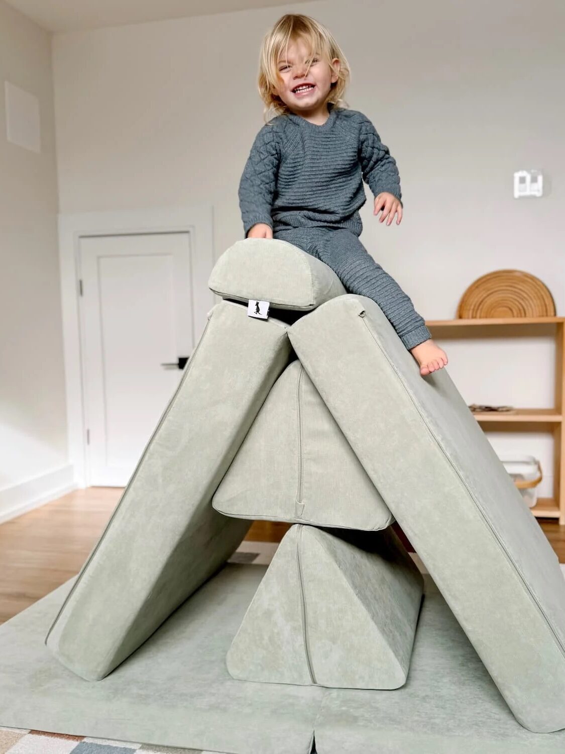 A young child in gray pajamas sits on top of a play couch structure made of soft, light gray foam cushions in a bright room.