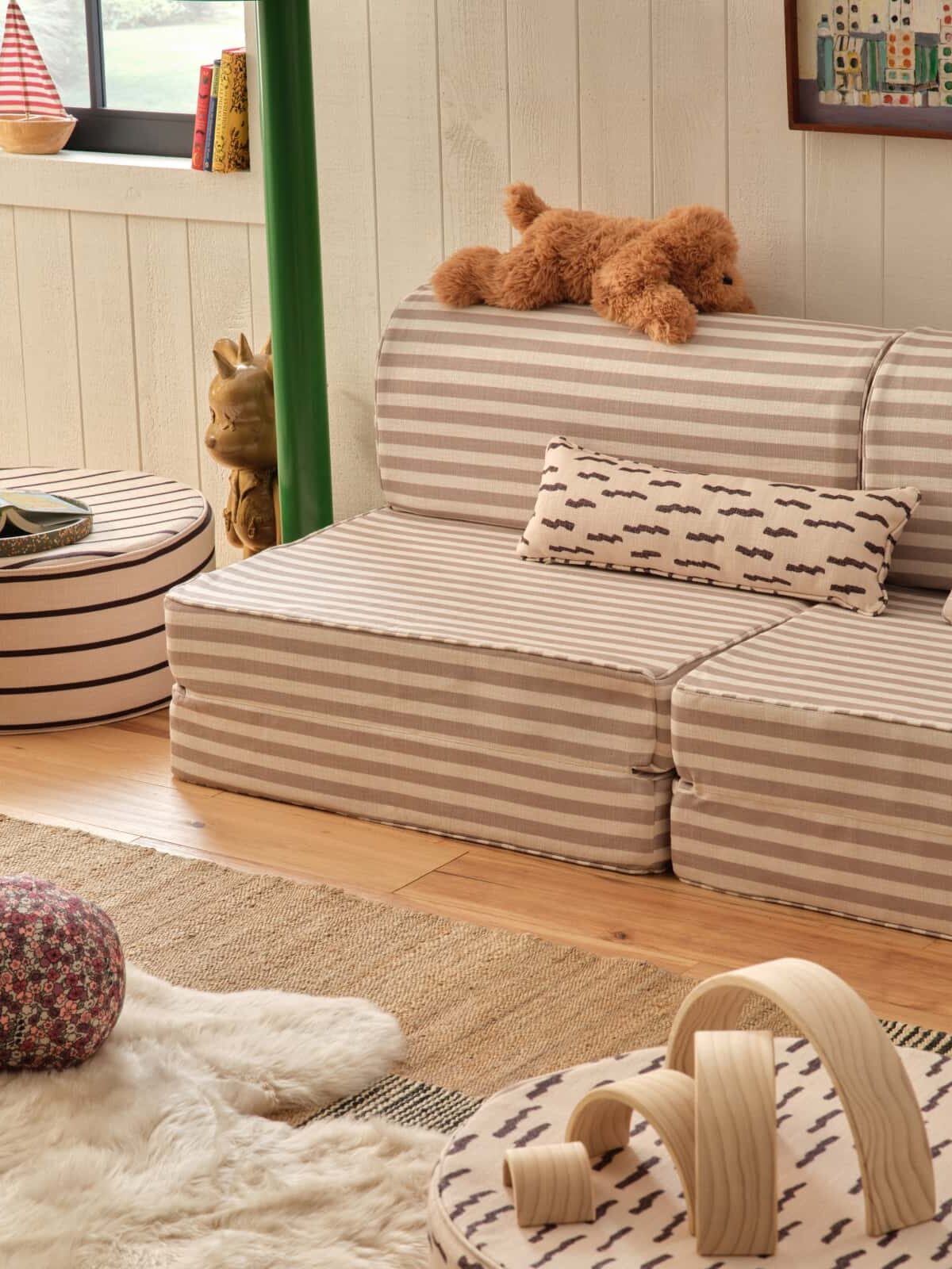 A cozy children's playroom with a striped fold-out sofa, plush toys, a round ottoman, a patterned pillow, and a soft rug on a wooden floor.