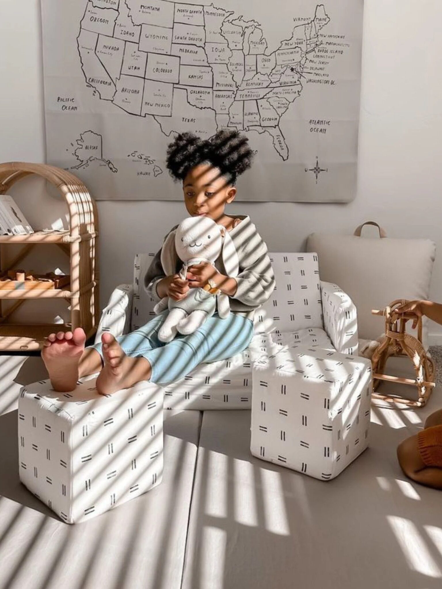 Two young children play in a sunlit room with patterned furniture; one sits on a sofa holding a plush toy, while the other stands nearby. A map of the U.S. hangs on the wall behind them.