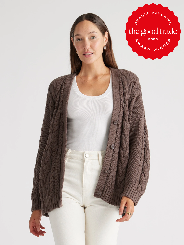A woman wearing a brown cable-knit cardigan over a white top and white pants stands against a plain background. A red "The Good Trade Award Winner 2025" badge is in the top right corner.