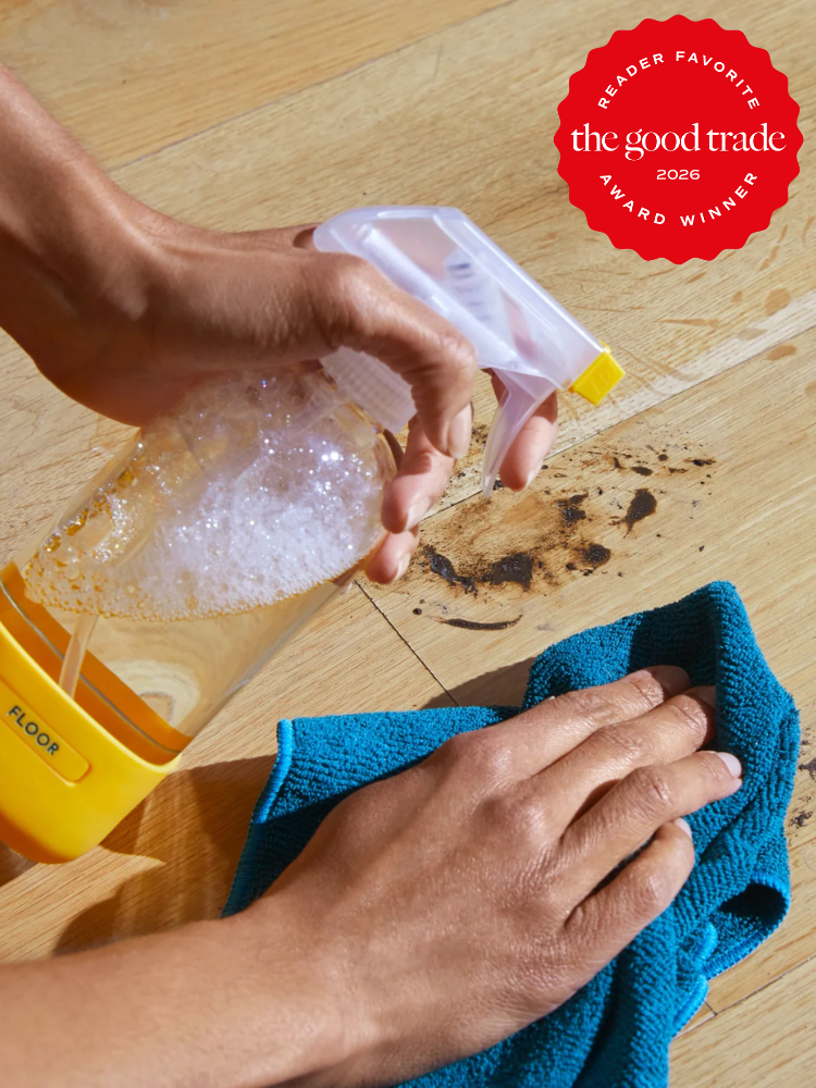 A person sprays cleaner from a labeled bottle onto a wood floor and wipes a dirty spot with a blue cloth. A red "The Good Trade Award Winner 2026" badge appears in the corner.