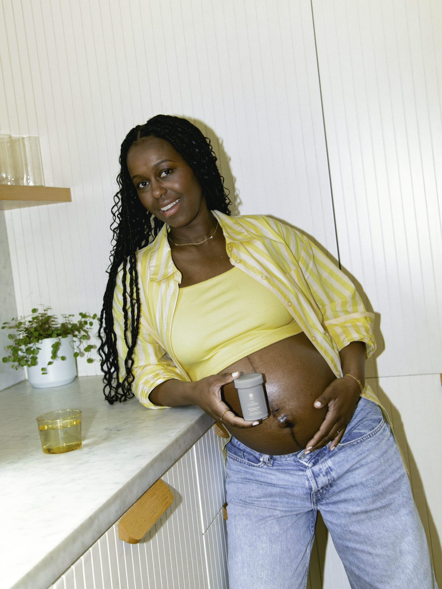 A pregnant woman in a yellow top and striped shirt leans against a kitchen counter, holding a small jar against her bare belly.